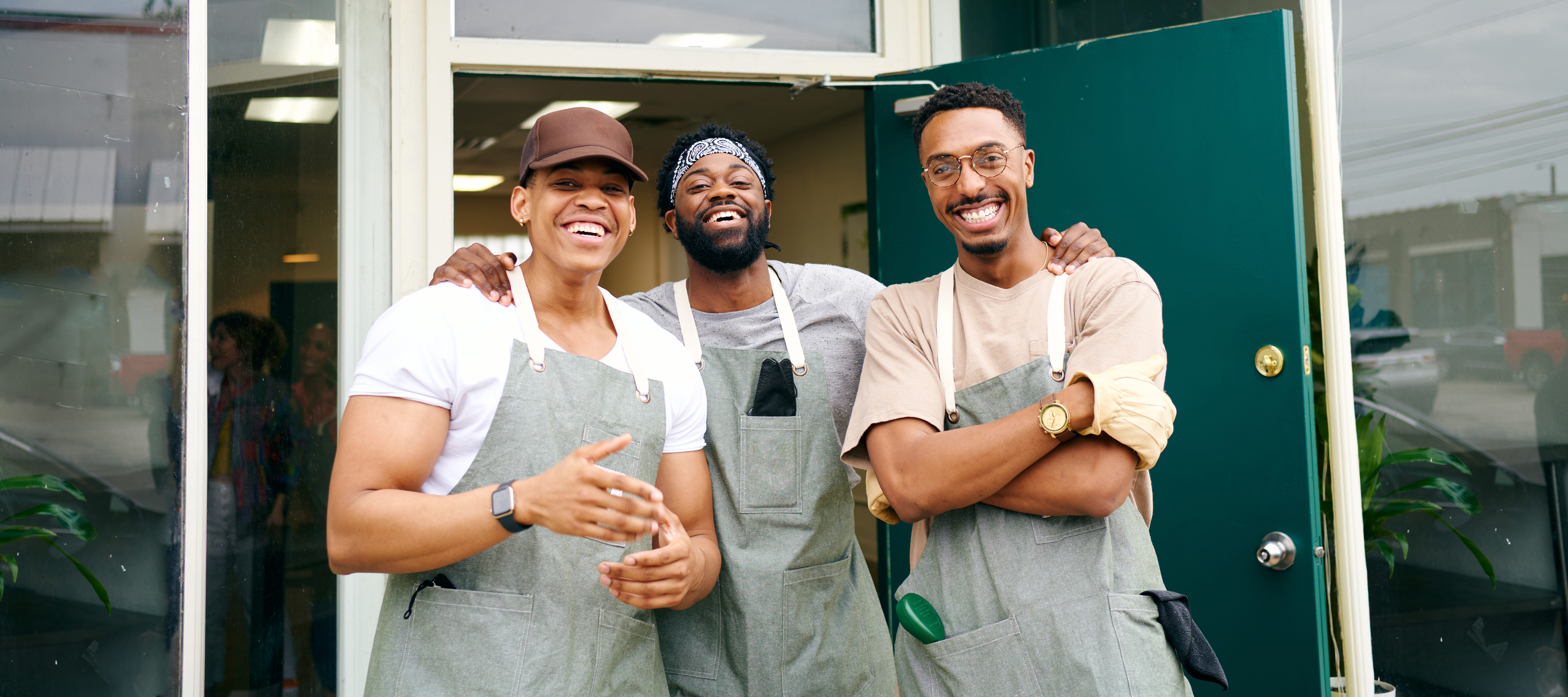 صورة لمجموعة ذكور من عمال الأعمال الصغيرة السود السعداء يضحكون على باب مشتل نباتات حضري يرتدون مآزر في دالاس